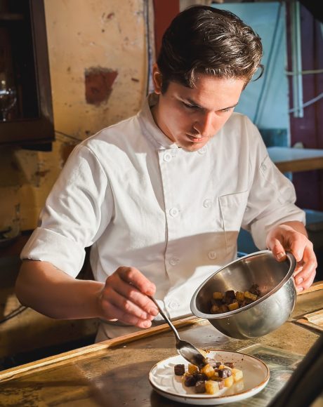 Chef in white clothing, plating a dish with potatoes and beef. on a counter.